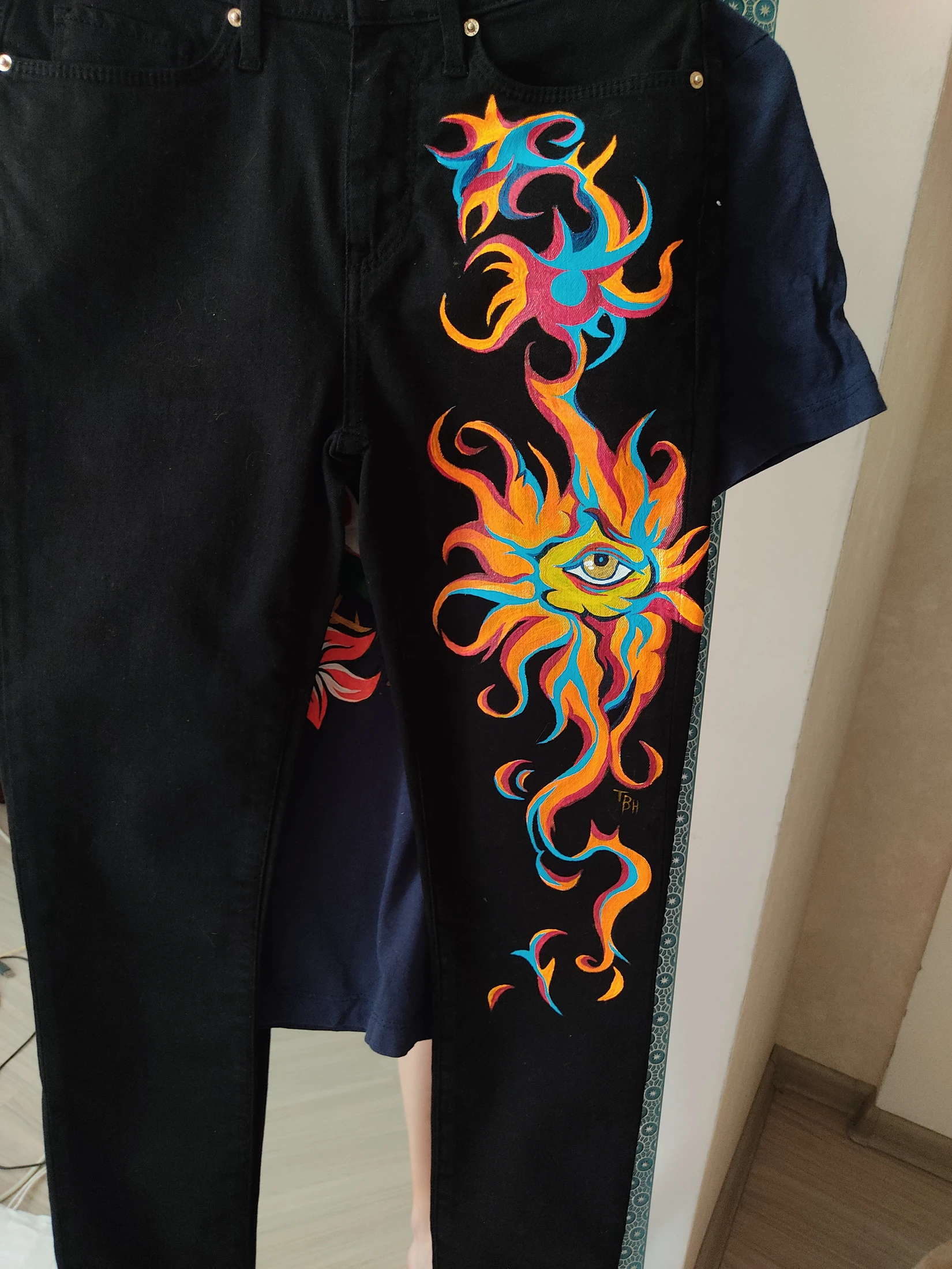 A single eye within orange, blue and pink flames extending down one black leg. Photograph of the hand-painted piece.