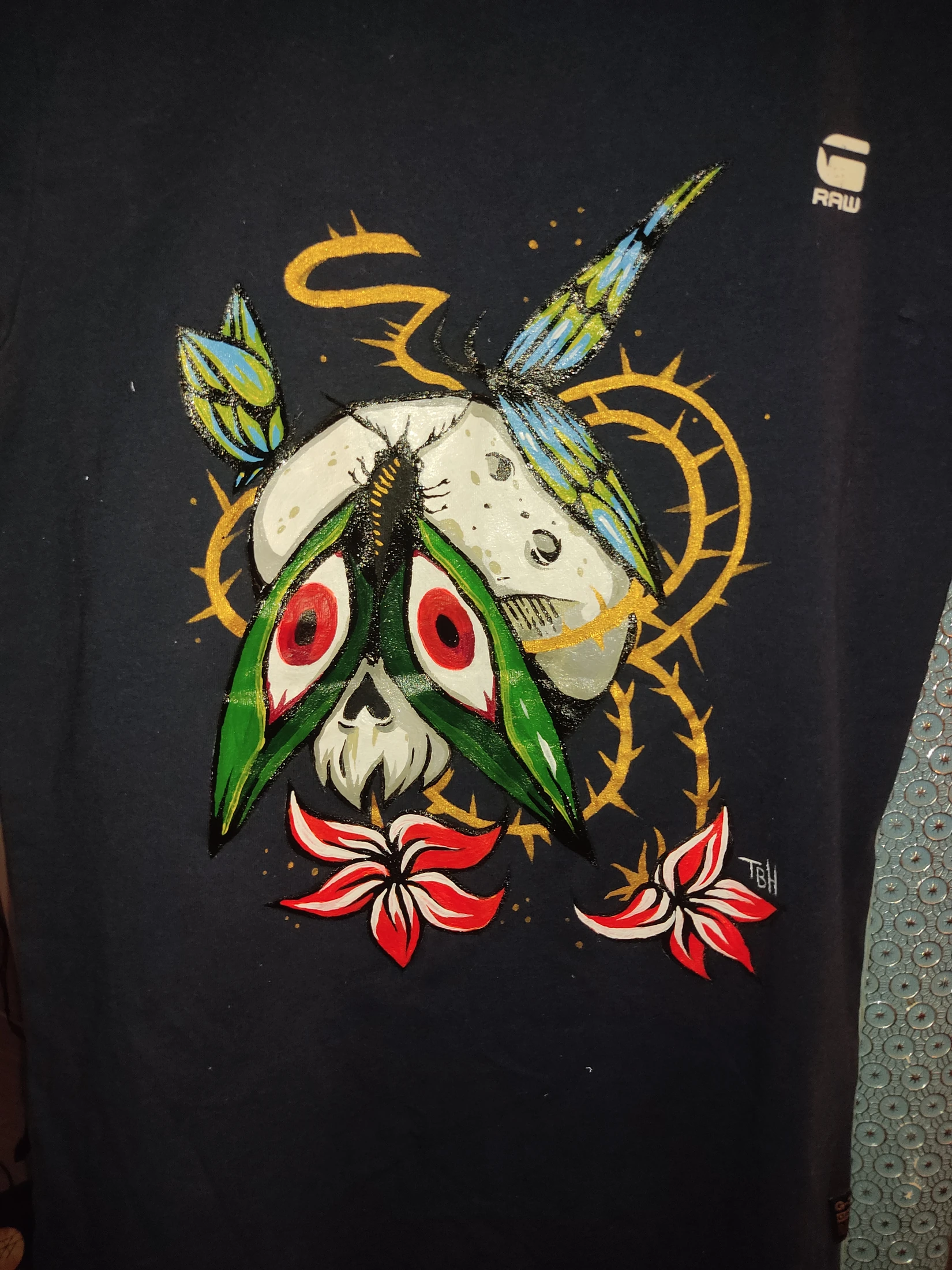 A skull-like moth beneath a pale moon, with green and yellow wings, gold curves and red-and-white flowers. Photograph of the hand-painted piece.