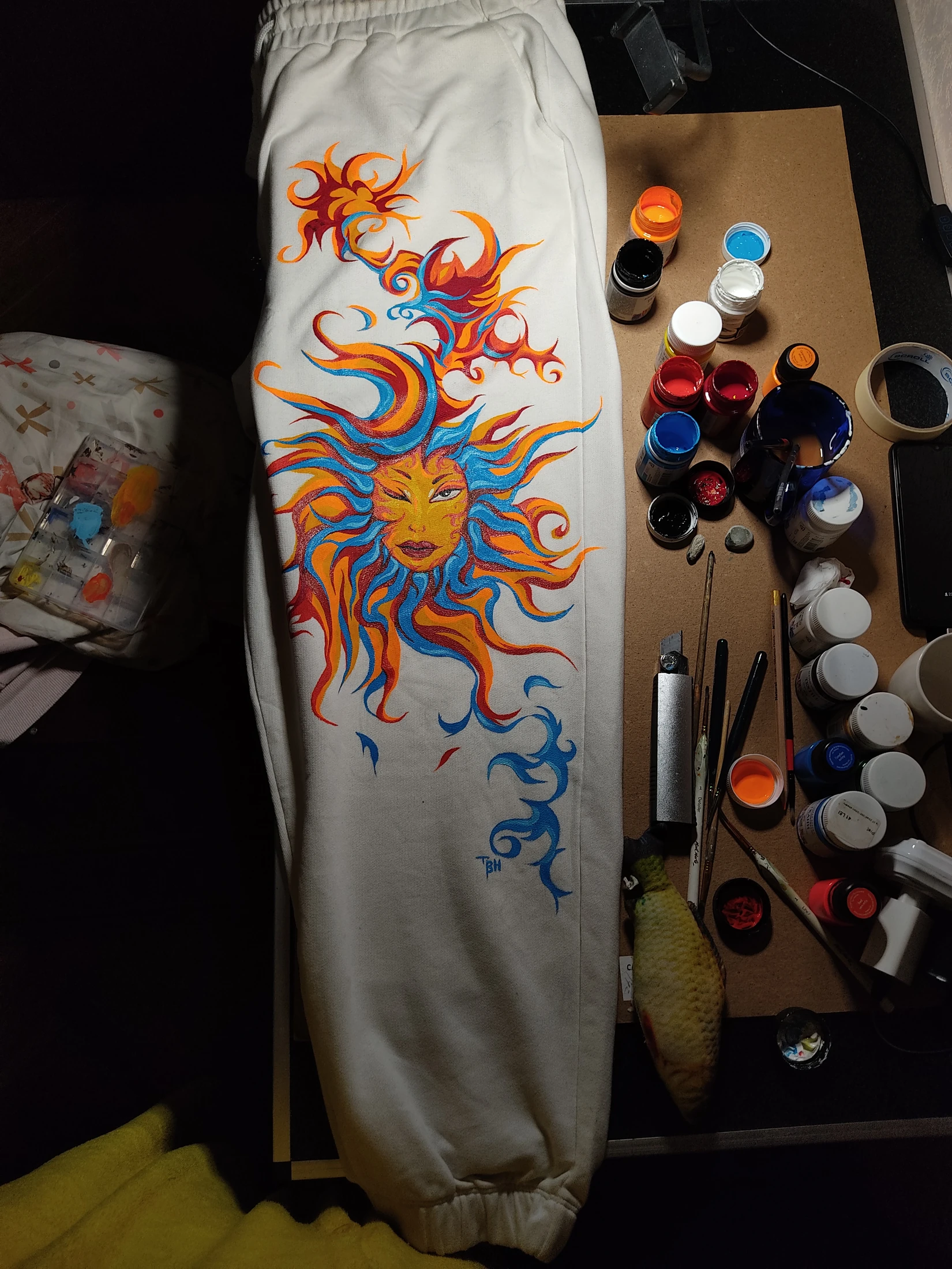 An orange sun face with flowing blue and red rays across a light trouser leg. Photograph of the hand-painted piece.