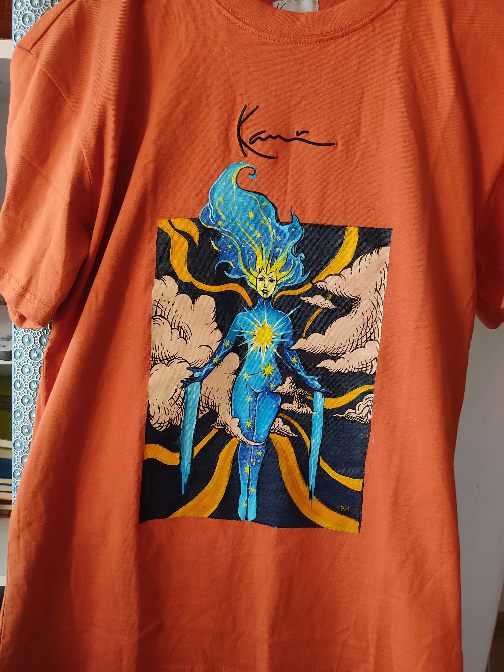 A blue figure with star motifs and flowing flame-like hair, framed by pale clouds and yellow curves. Photograph of the hand-painted piece.